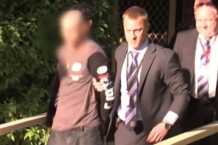 A man in brown top and pants being led away by three police officers. His face is obscured for legal reasons