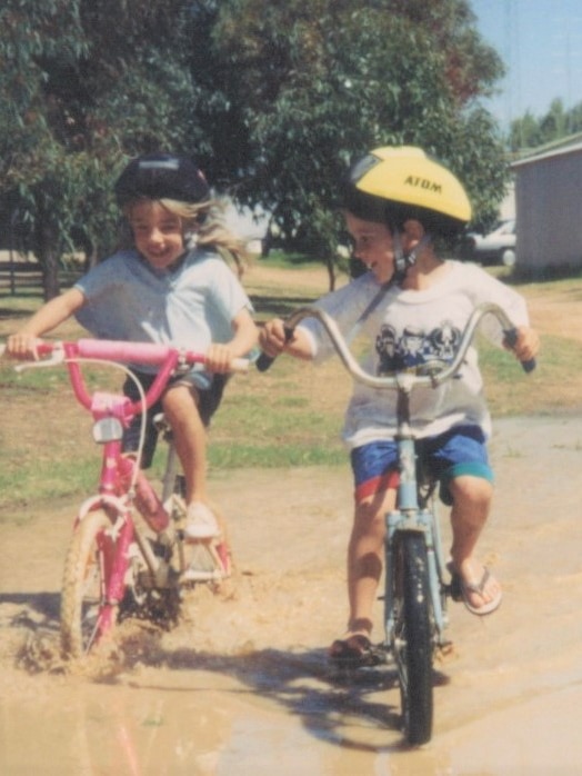 An old film photo of a young girl and boy wearing helmets, biking through a muddle puddle on a sunny day.