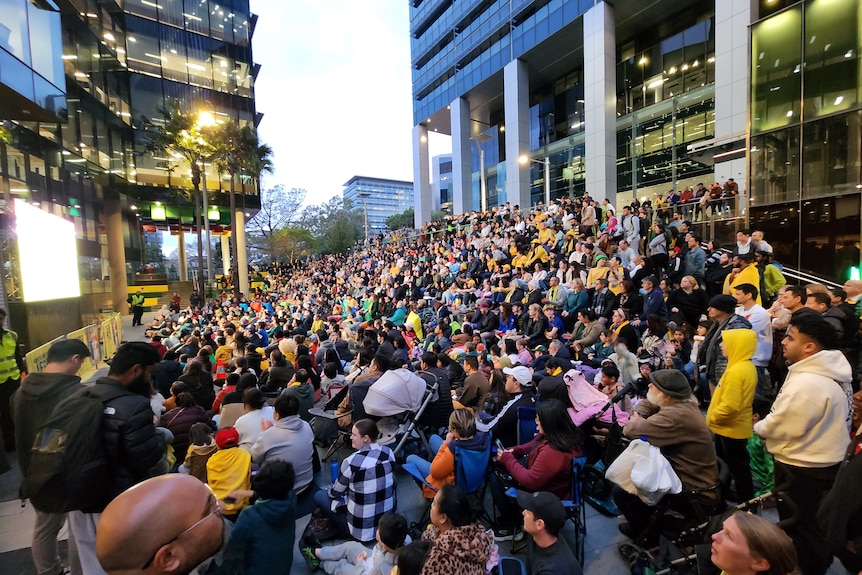 A crowd of football fans wearing yellow watch a television screen outdoors