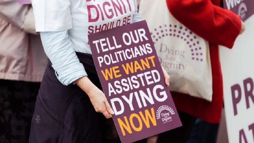 Woman holding a sign that says "Tell our politicians we want assisted dying now"