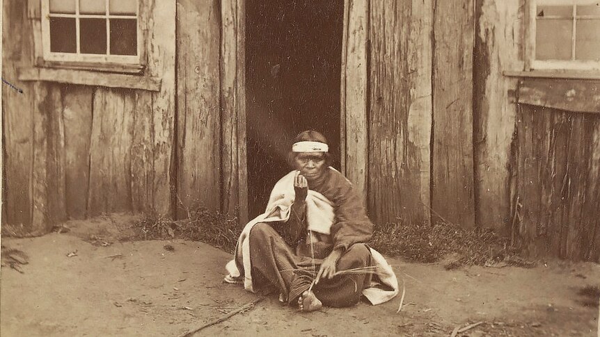 A Gilgar Gunditj woman sits weaving in front of a bark shack. She wears blankets and a head band.