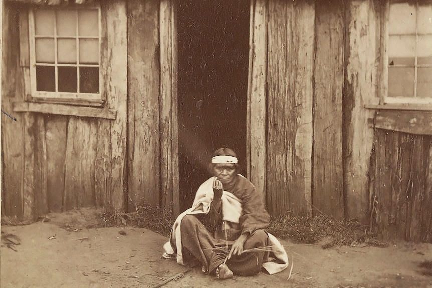 A Gilgar Gunditj woman sits weaving in front of a bark shack. She wears blankets and a head band.