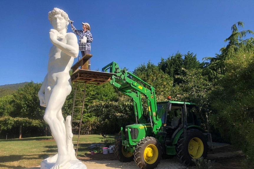 A person applies a paint brush to the head of a large replica of Michelangelo's David