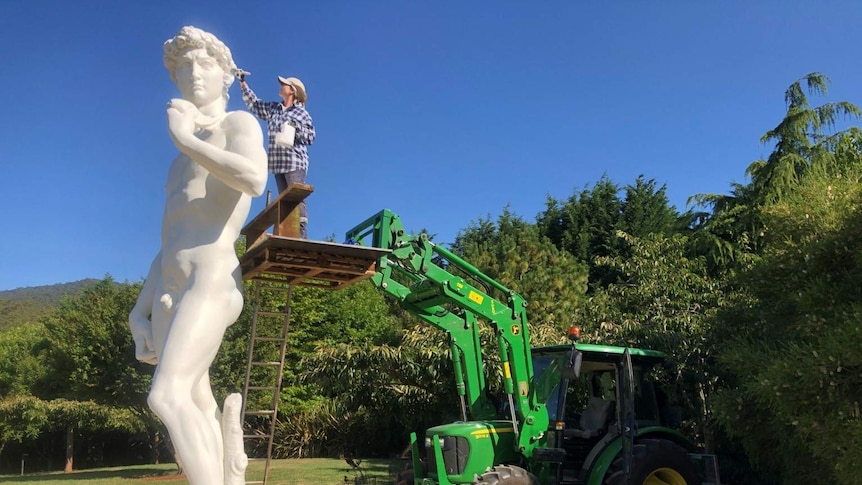 A person applies a paint brush to the head of a large replica of Michelangelo's David