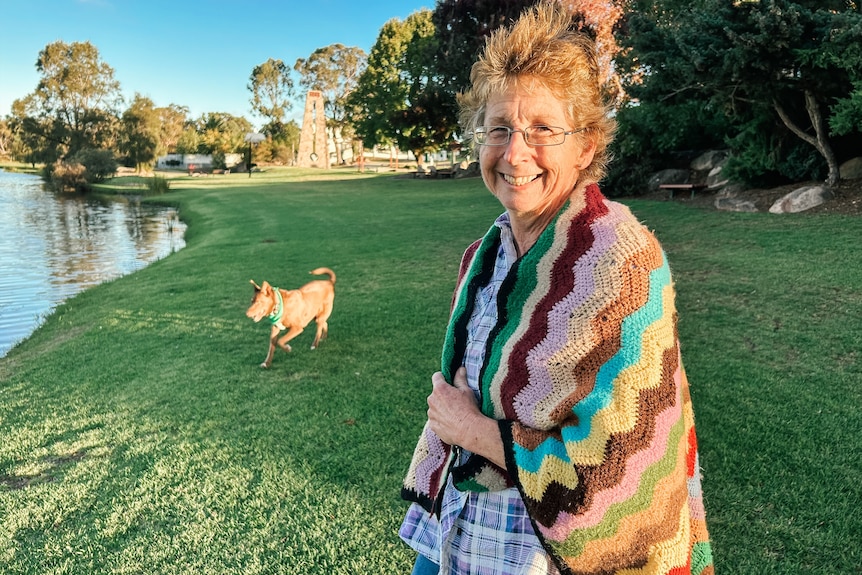 A woman with a blanket wrapped around her shoulders smiles in a park with a dog in the background