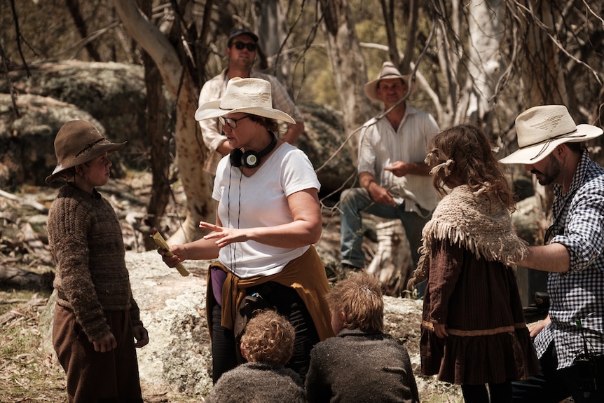 A female director stands outdoors wearing a wide brim hat among a group of cast and crew