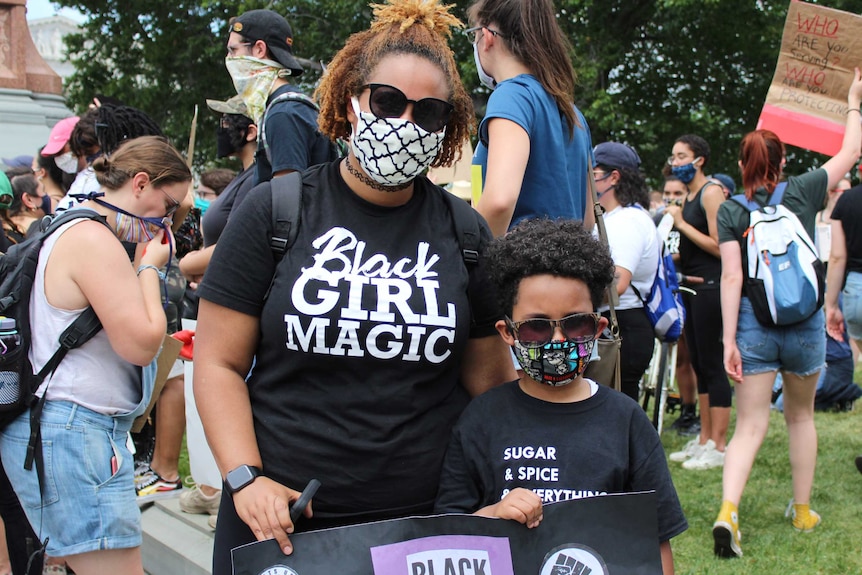 A woman in a black girl magic t-shirt and face mask stands with her son