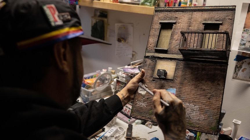 A man dressed in streetwear holds a paintbrush against a miniature model of an urban brick wall