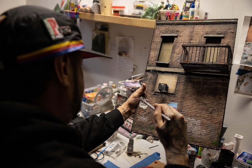 A man dressed in streetwear holds a paintbrush against a miniature model of an urban brick wall