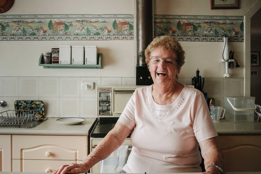 An elderly woman sits at her kitchen bench