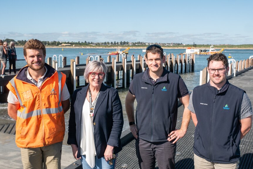 One woman and three men, two wearing blue sleeveless jackets with council logo, stand in front of a boat ramp, smiling.