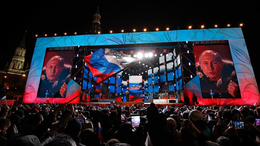 View from a packed crowd at night, outside, looking towardws Putin on stage with two giant screens showing putin's face