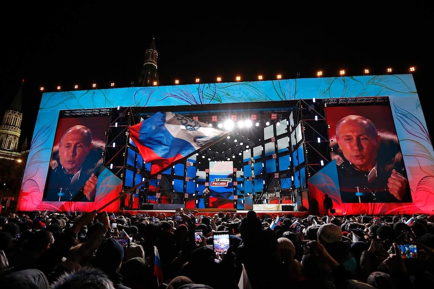 View from a packed crowd at night, outside, looking towardws Putin on stage with two giant screens showing putin's face