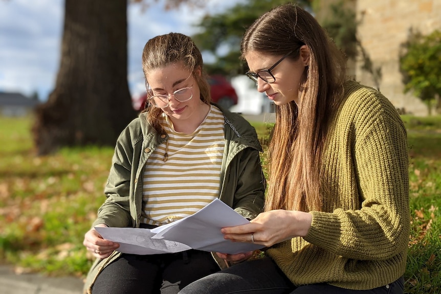 Two young women wearing glasses sit next to each other and read a document