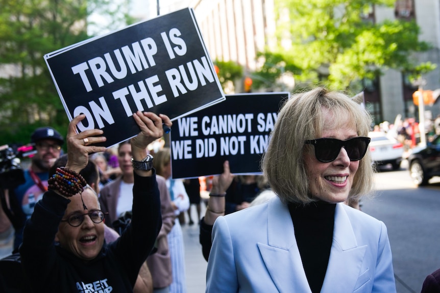 A woman with a blonde bob and sunglasses smiles as she walks past protesters holding up signs saying Trump is on the Run