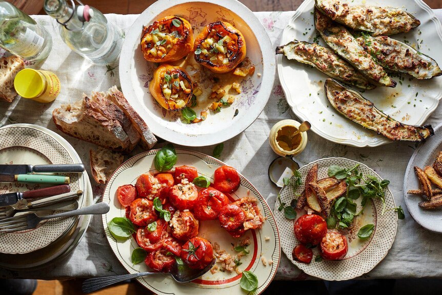 A dining table covered with plates of food: roast tomatoes, stuffed capsicums and zucchini
