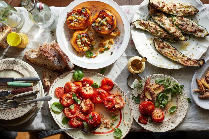 A dining table covered with plates of food: roast tomatoes, stuffed capsicums and zucchini