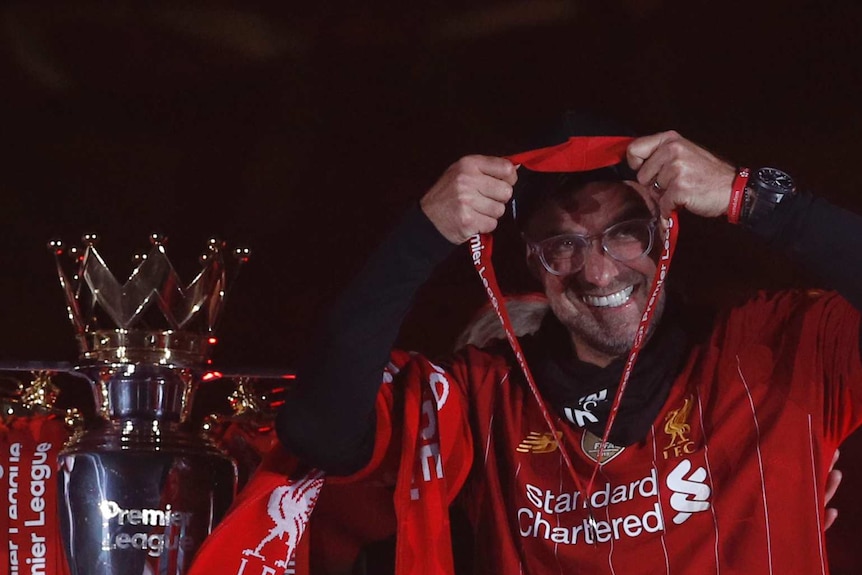 A grinning manager holds the winner's medal up, while standing next to the Premier League trophy.