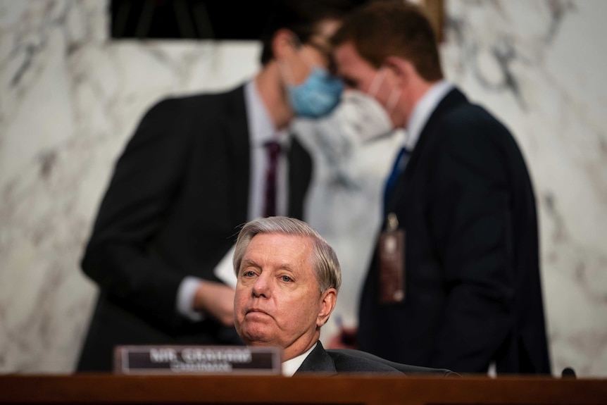 A man sits behind a desk as he listens to a hearing, while two men wearing face masks can be seen whispering behind him.