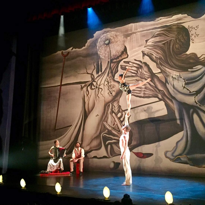 A female acrobat balances with one leg on the out-stretched arm of a man on stage.