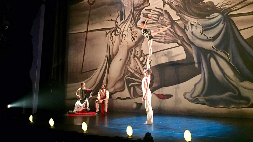 A female acrobat balances with one leg on the out-stretched arm of a man on stage.