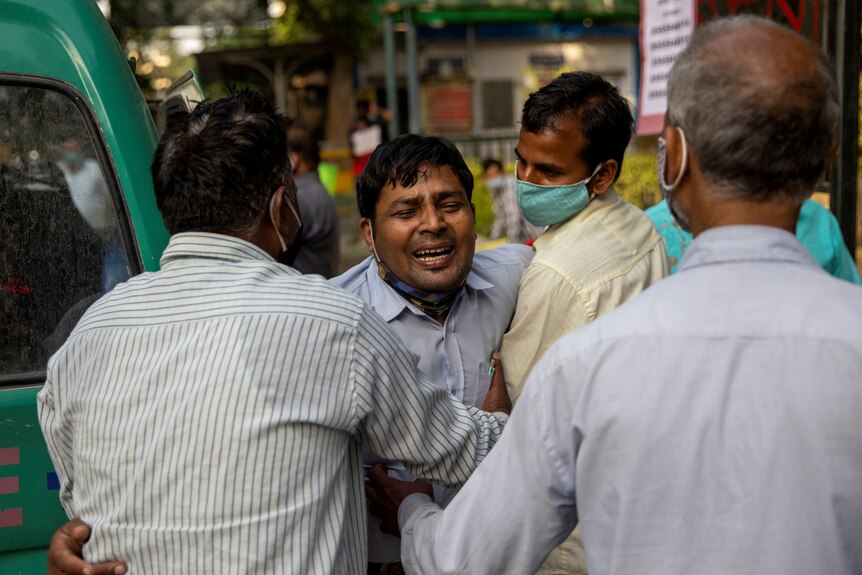An Indian man cries while held back by other men wearing face masks 