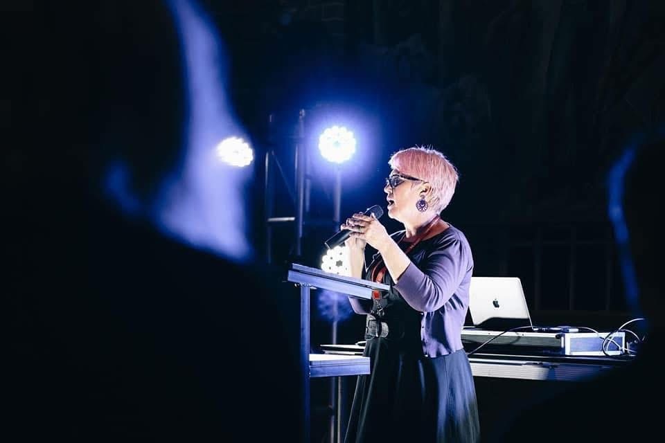 A woman with short pink hair stands on a stage, speaking into a microphone, bright lights behind her