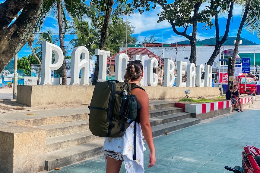 Kate carries her carry-on backpack, which is fairly small, by a large sign reading: Patong Beach.