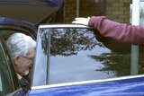 Rolf Harris exits a blue car outside his home in Berkshire, while a man in a maroon hoodie stands nearby.