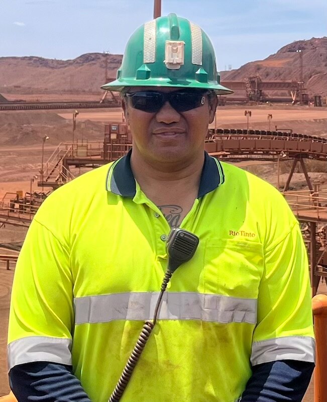 A man in a bright green t-shirt wears hard hat, sunglasses, has a radio clipped to his shirt, tattoo can be seen on chest.