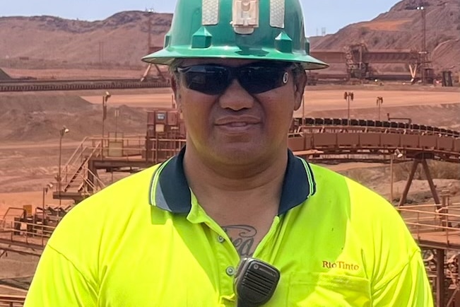 A man in a bright green t-shirt wears hard hat, sunglasses, has a radio clipped to his shirt, tattoo can be seen on chest.