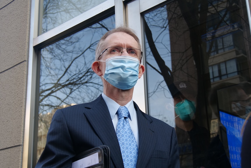 A man in a suit and blue face mask, with a leather folder tucked under his arm