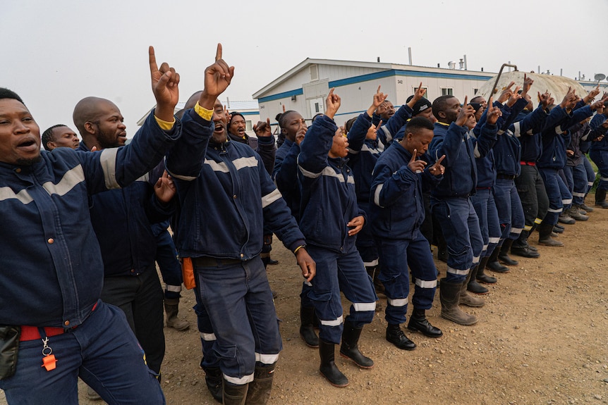 A group of South African firefighters in navy uniforms are captured mid-dance, hands in the air, fingers pointing to the sky.