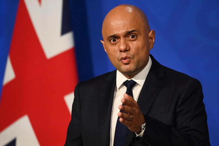 A man in a suit in front of UK flag looks at the camera and gestures with hand 
