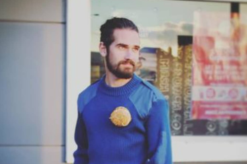 Young man with a beard and man bun wearing a blue woollen jumper with a hamburger brooch