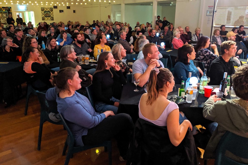 A wide shot showing a big crowd of people sitting in a community hall looking to the right of the frame.