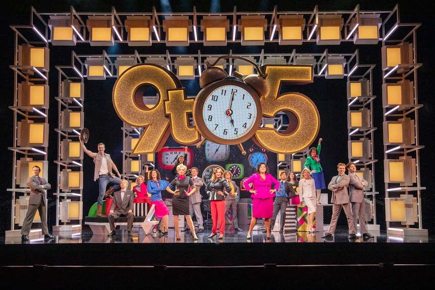 A giant gold 9 to five clock and numerous people in colourful outfits on a large brightly-lit stage.