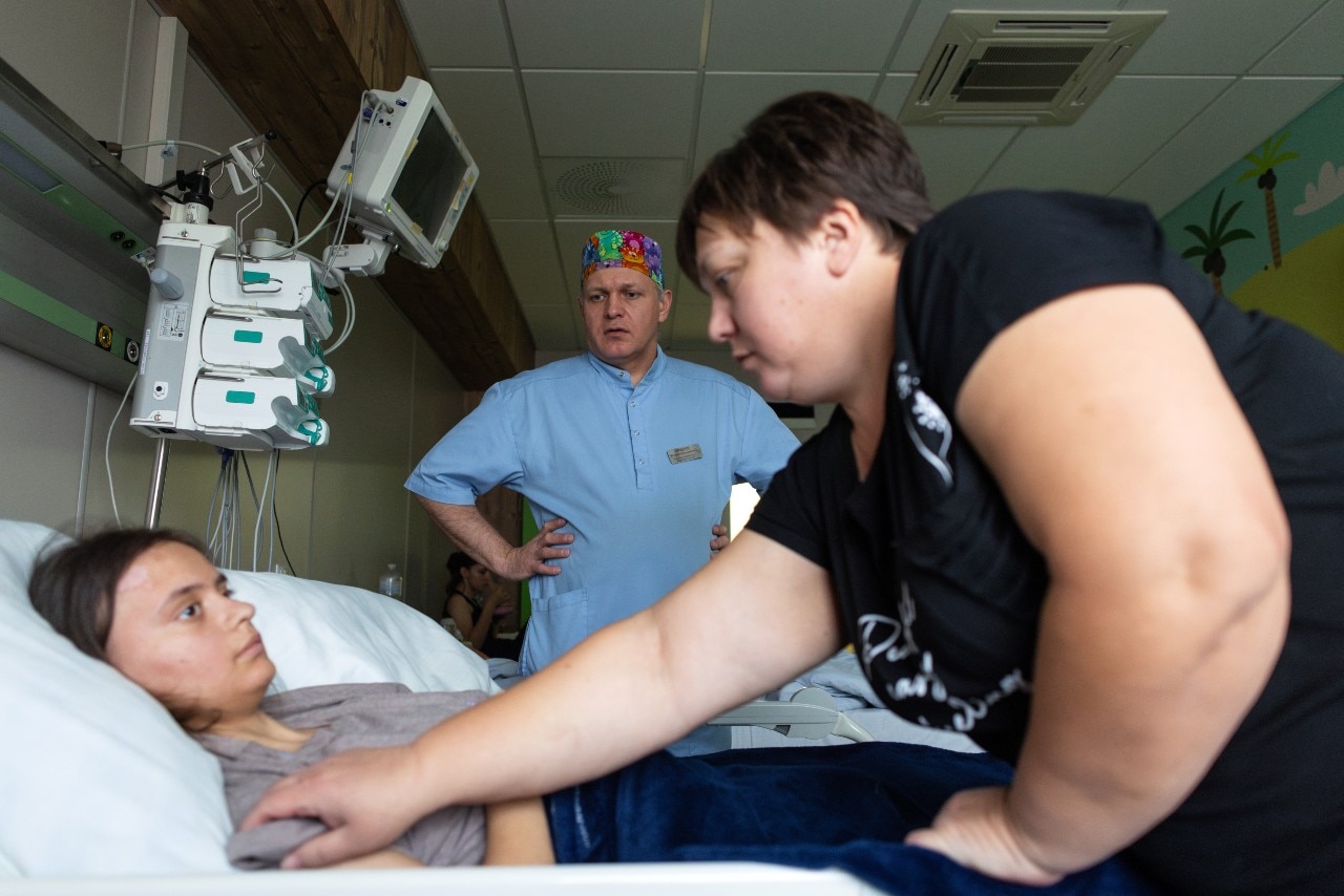 A girl in a bed looking at a woman who is leaning over and touching her. A man is in the background looking on