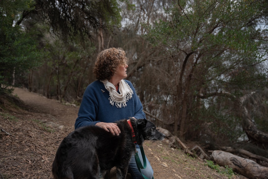 A woman in blue jumper and white scarf crouches down with her black dog with white facial hairs.