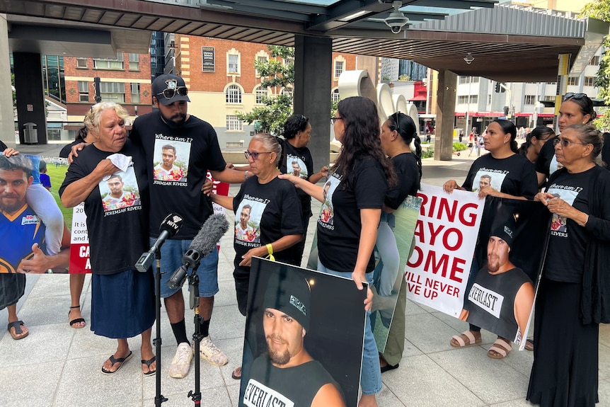 A group of people hold up pictures of Jeremiah Rivers while standing in front of microphones