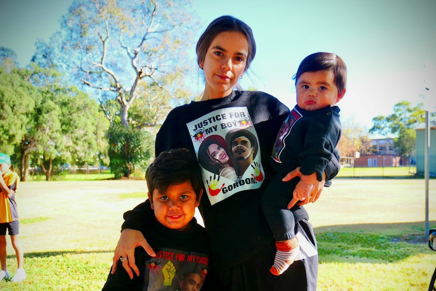 Жена стои с малко дете и малко момче, облечена в черен пуловер със снимка на себе си и на мъж във формата на сърце.