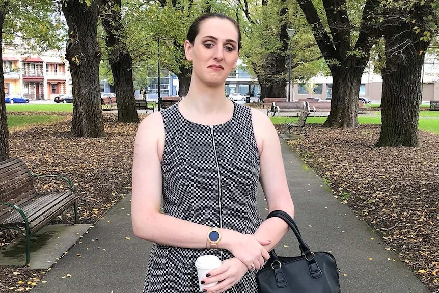 Brooke stands in a park wearing a grey dress and holding a handbag.