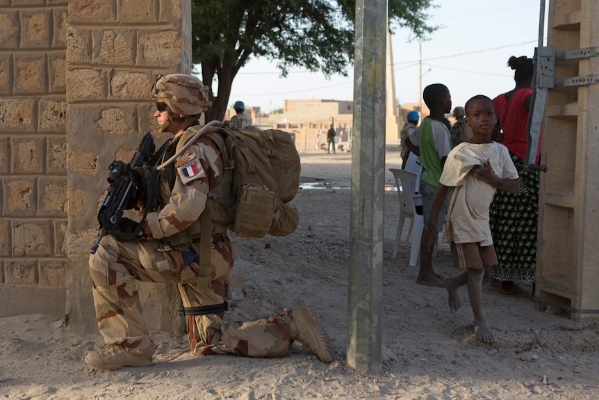 A French soldier holding a gun kneels as a young Malian boy walks by.
