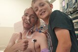 Patient Joshua Leveridge in a hospital bed, with his 10-year-old son Reuben standing alongside, with their heads together.