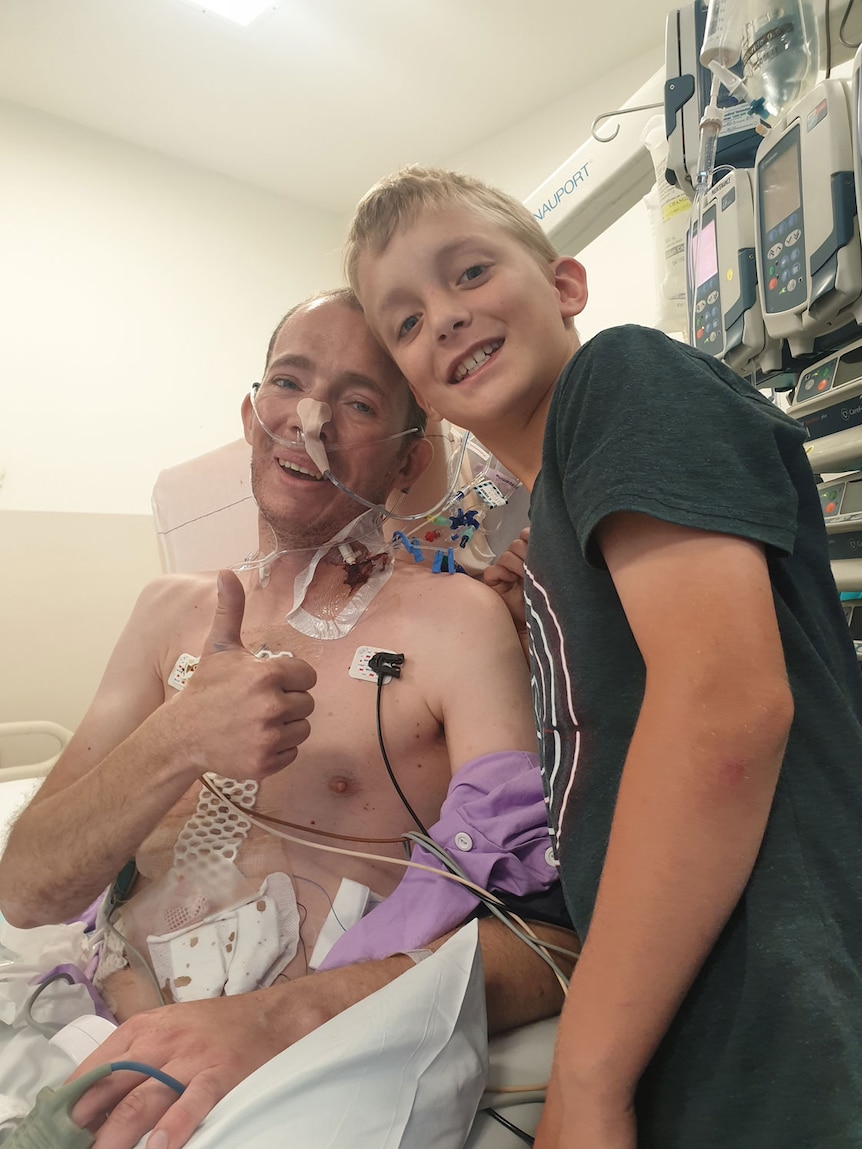 Patient Joshua Leveridge in a hospital bed, with his 10-year-old son Reuben standing alongside, with their heads together.