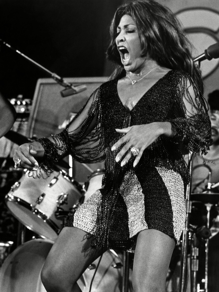 A black and white photo of a Black woman, Tina Turner, dancing on stage in the 70s