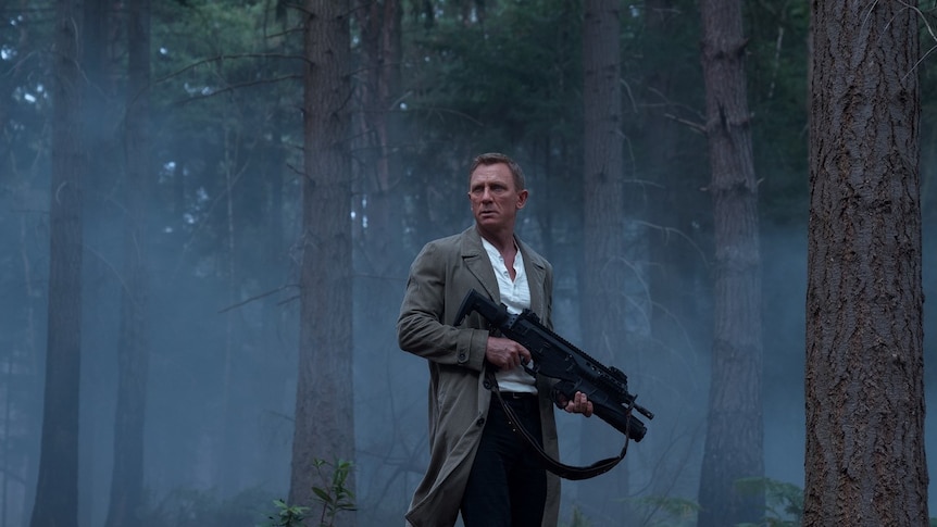 A still from the Bond film No Time To Die showing Bond in a misty pine forest holding a large black gun