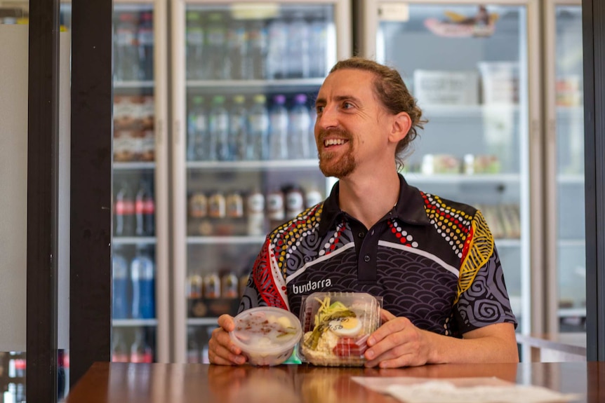 A man wearing an Indigenous-design polo shirt smiles off camera holding two salads.