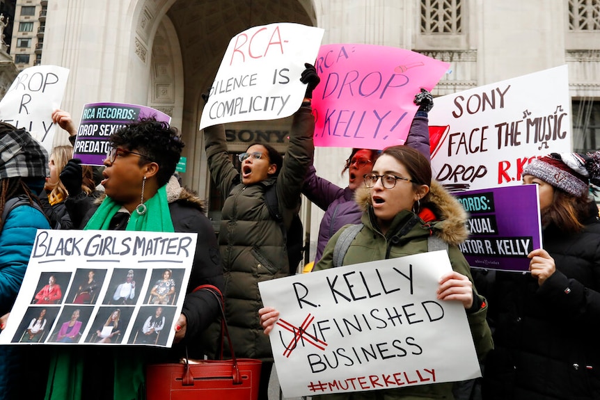 Women dressed in winter jackets holding handwritten signs with slogans such as "mute R Kelly"