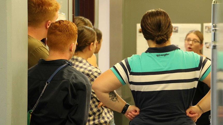 Six students stand in a crowded room with their backs to the camera as a teacher explains coffee making techniques to them.